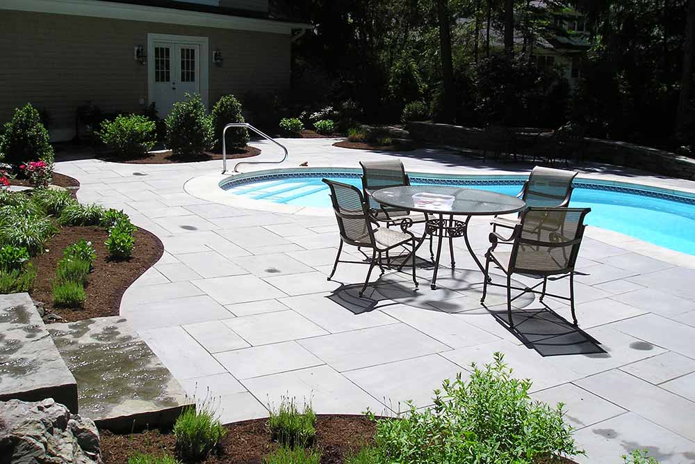inground pool with stone deck, table and chairs and greenery